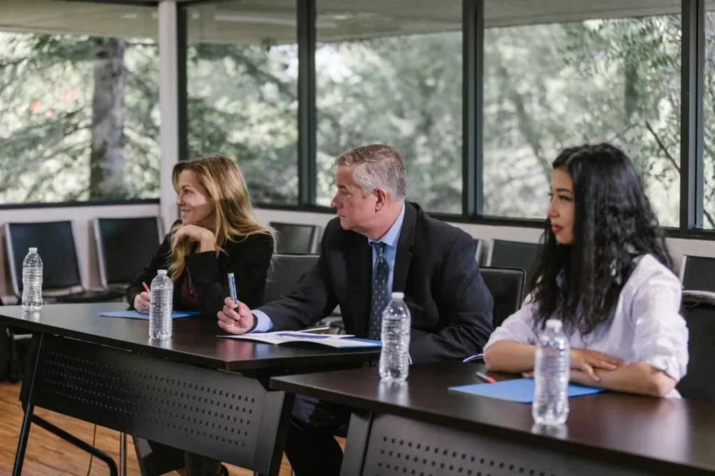 Three people seated at conference tables with notebooks, pens and water bottles during a meeting.
