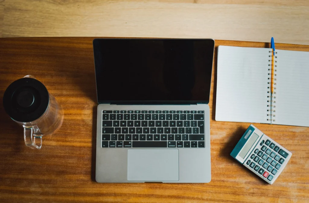 Overhead view of a laptop, calculator, blank notebook and coffee mug on a wooden desk.