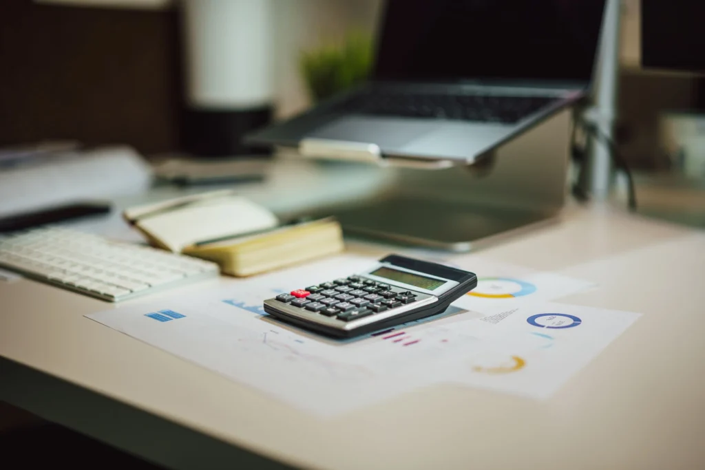 A calculator on printed financial charts beside a keyboard, notebook and laptop.