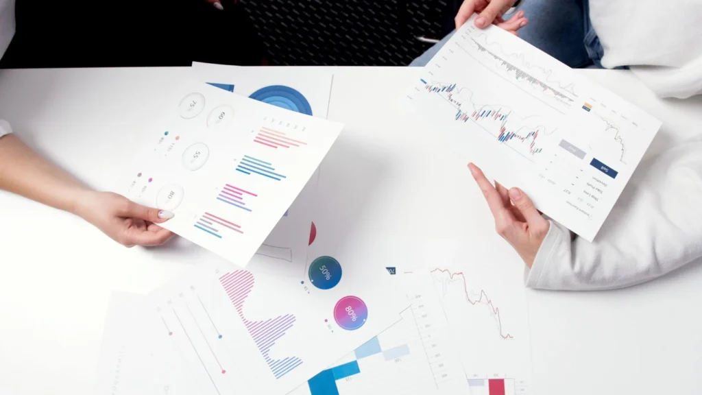 Two people review printed investment charts and graphs across a desk.