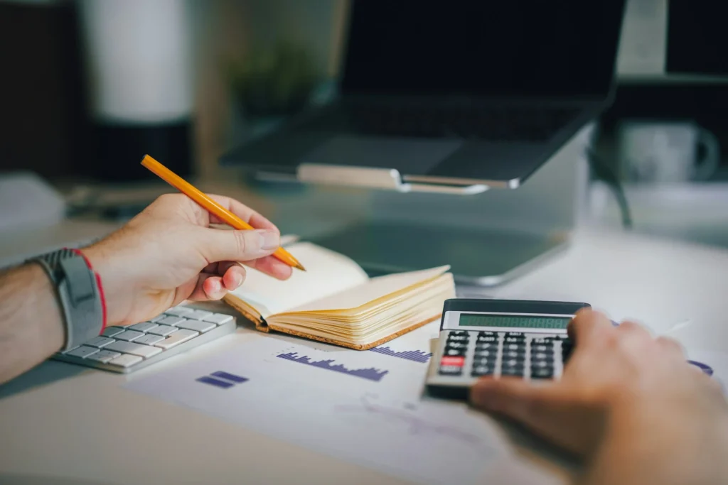 Hands using a calculator and pencil beside a notebook and printed financial charts.