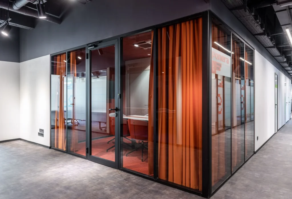 Glass-walled office meeting room with red chairs and curtains beside a bright corridor.